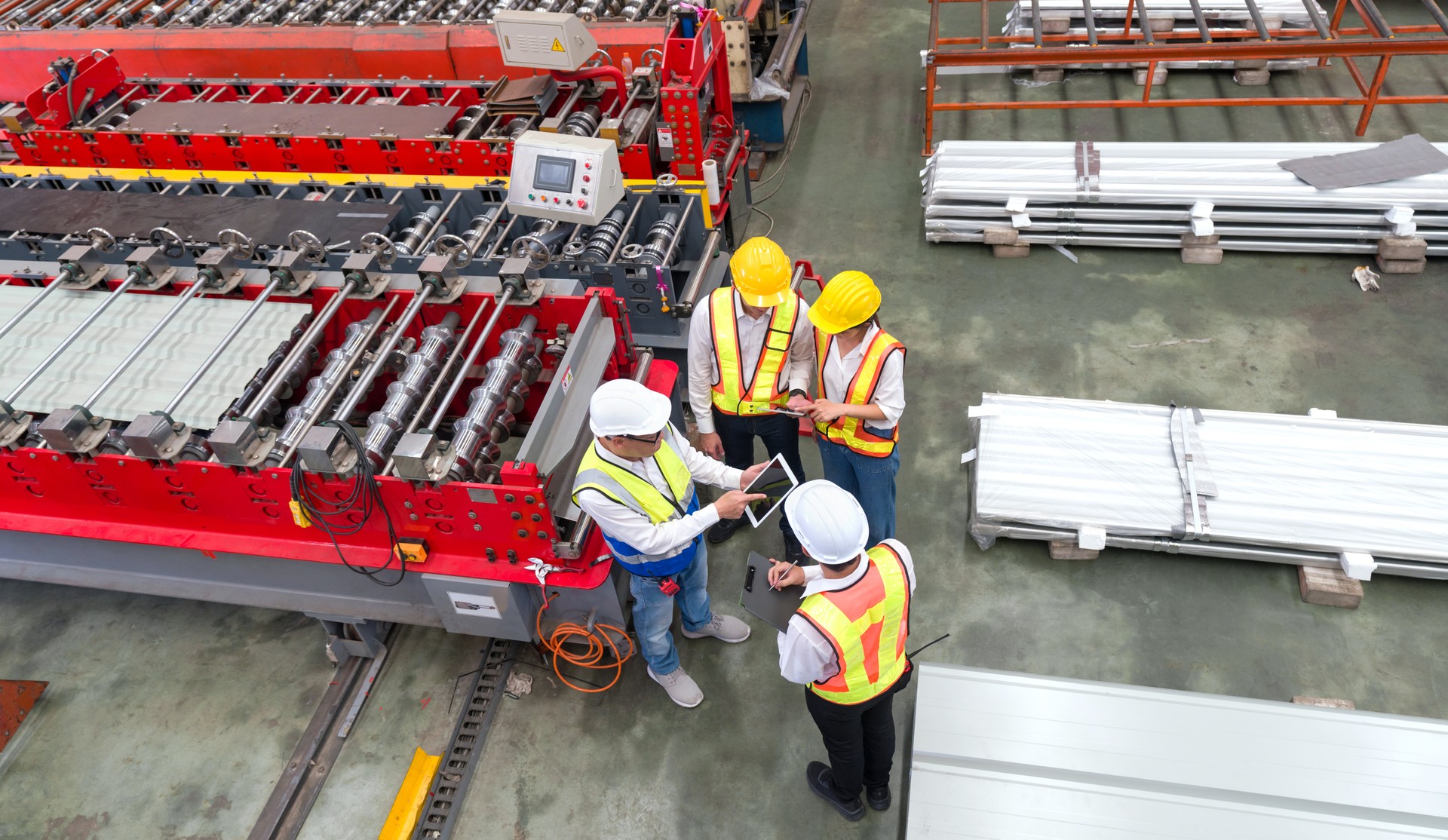 Senior engineer manager trains new employees within the metal sheet factory. Everyone wear safety vest and hardhat. Large machines and metal sheets are in the working area.